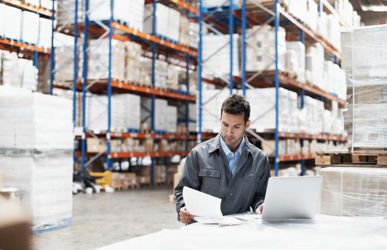 A man using EPR software on a laptop in a large warehouse, reviewing documents surrounded by shelves filled with boxes and pallets.