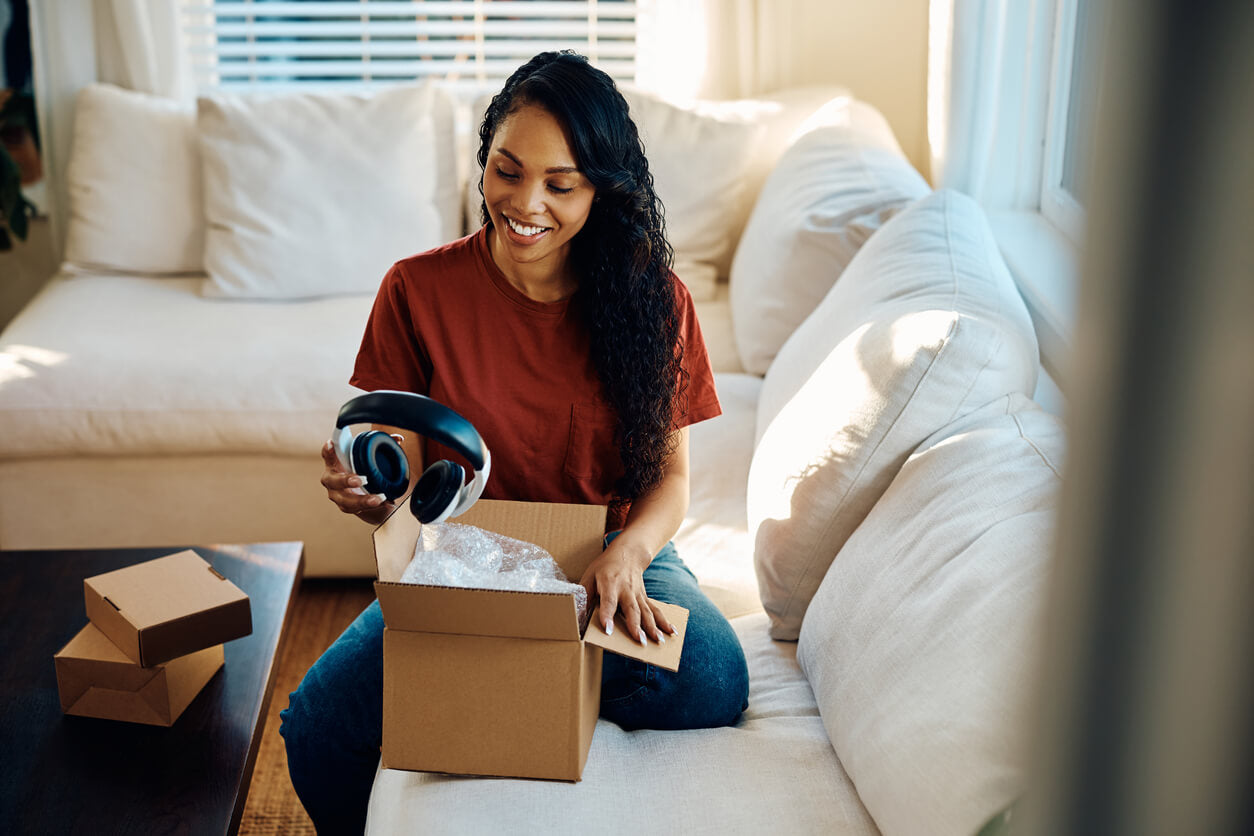 A happy woman sitting on a couch, unboxing a pair of headphones from a cardboard package, an item necessary to report with EPR software.