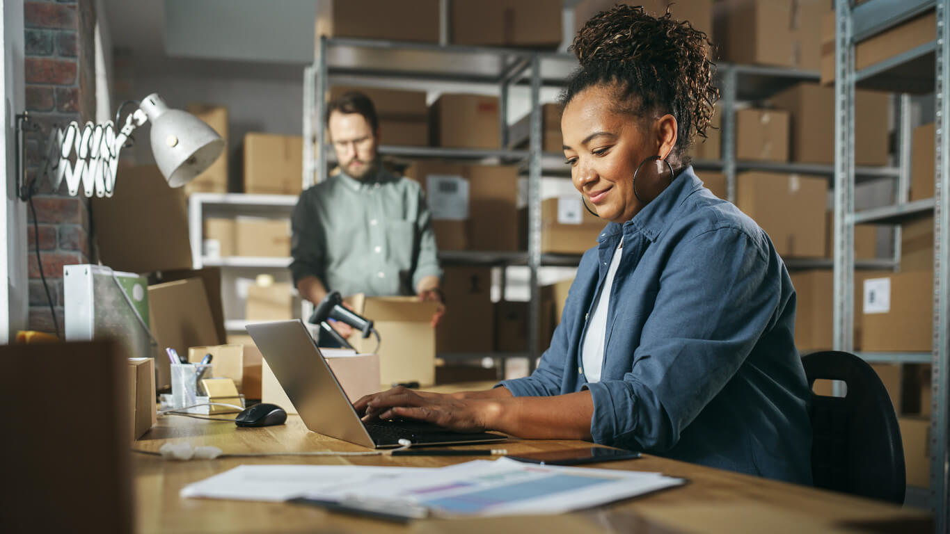 A smiling woman using EPR software on a laptop in a warehouse, with boxes and a colleague in the background managing inventory.